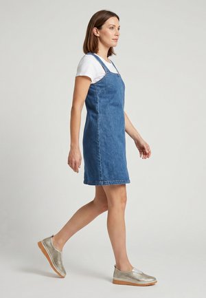 Vrouw lopend met een wit shirt met korte mouwen onder een blauwe spijkerjurk en metalen instapschoenen met rubberen zolen.