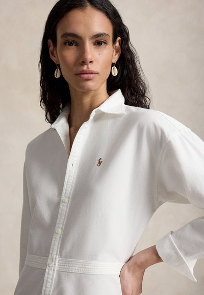 Γυναίκα με μακριά σκούρα μαλλιά που φοράει λευκό πουκάμισο με κουμπιά, με μικρό κεντητό λογότυπο και σκουλαρίκια σε σχήμα κοχυλιού, ποζάροντας με το χέρι στη μέση.