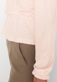 Blusa texturizada em rosa claro com mangas com babados e cintura drapeada, combinada com calças castanhas de corte reto com bolsos laterais.