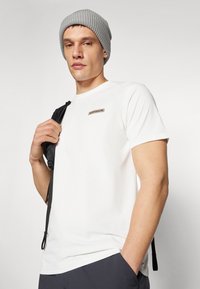 Tricou alb cu mânecă scurtă, dotat cu un mic patch logo maro, asortat cu o căciulă gri din tricot și o curea de rucsac neagră peste umăr.