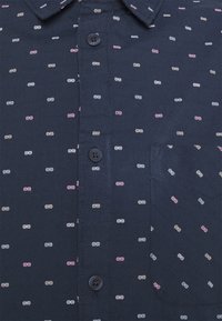 Námořnická košile na knoflíky s kapsou, zdobená vzorem malých barevných teček v růžové, bílé a světle modré barvě na hladké látce.