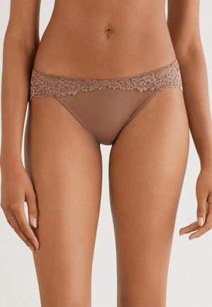 Mujer vistiendo ropa interior estilo bikini de encaje color marrón claro, mostrando la parte media del cuerpo y los muslos superiores contra un fondo neutro.