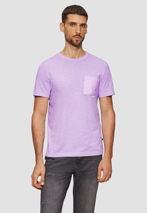 Pacte paarse T-shirt met korte mouwen van zachte stof, met een zak op de voorkant. Draagt met donkergrijze versleten jeans.
