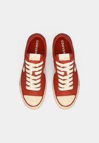 Sneakers in tela rossa con lacci crema, punta in gomma e linguetta beige con il logo "Converse All Star". Suole piatte e dettagli laterali.