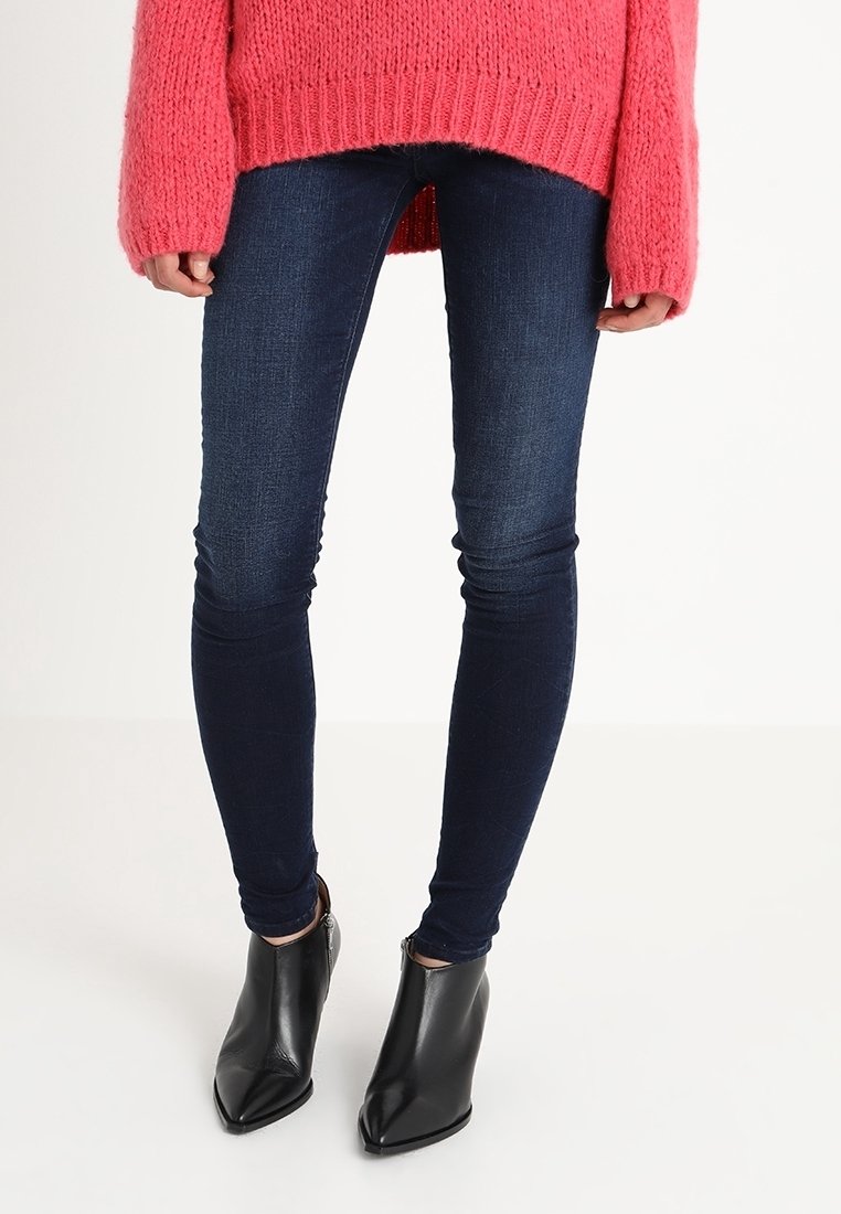 Jeans ajustados de mezclilla azul combinados con un suéter de punto suelto de color rosa brillante y botas de tobillo negras de punta afilada con textura brillante.
