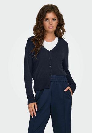 Jonge vrouw met lang golvend haar draagt een marineblauwe knopen cardigan over een wit shirt en marineblauwe broek, poseert met een hand in haar zak.