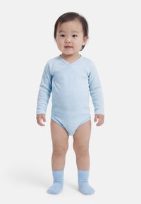 Νήπιο που στέκεται, φοράει ένα ανοιχτό μπλε ριγωτό κορμάκι με μακριά μανίκια και ταιριαστά μπλε καλτσάκια, μπροστά σε απλό λευκό φόντο.