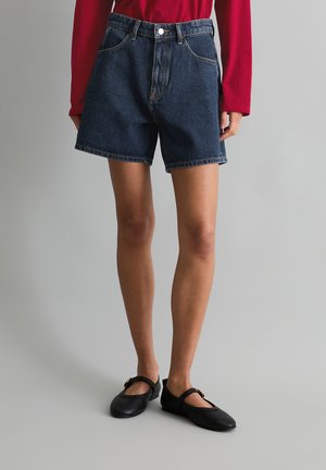 Persoon draagt donkerblauwe denim shorts, rode longsleeve top en zwarte Mary Jane schoenen, staand tegen een effen lichtgrijze achtergrond.
