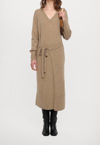 Robe en maille beige avec un col en V, des manches longues et une ceinture à nouer à la taille. Présente une silhouette décontractée et un ourlet arrivant aux genoux. Portée avec des bottes noires.