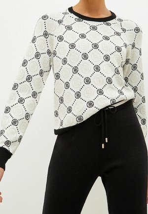 Pull blanc avec un motif circulaire noir et gris, doté de poignets et d'ourlet côtelés noirs, porté avec un pantalon noir. Texture en maille légère.