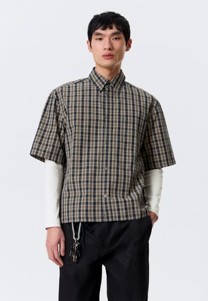 Young man wearing a checkered short-sleeve button-up shirt over a white long-sleeve shirt, black pants, holding keys attached to a carabiner.