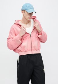 Ροζ cropped hoodie με ξεφτισμένες άκρες και φερμουάρ μπροστά, σε συνδυασμό με μαύρο παντελόνι φόρμας με λωρίδες στις πλευρές σε λευκό χρώμα, φορεμένα με ανοιχτό μπλε καπέλο.