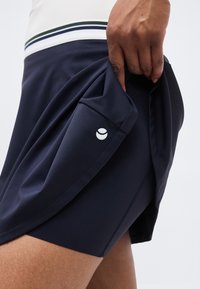 Hånd løfter kanten på navy treningsskjørt, som avslører innebygde shorts med liten hvit logo på siden.