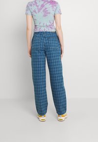 Osoba ubrana w niebieskie spodnie z wysokim stanem w diamentowy wzór oraz krótki rękaw w fioletowo-biały wzór tie-dye, stojąca na tle bez wzorów.