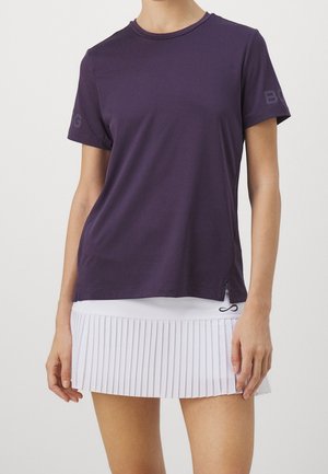 Person trägt ein dunkelviolettes, kurzärmliges Hemd und einen weißen Faltenrock mit einem kleinen schwarzen Logo nahe dem Saum.