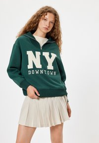 Sudadera verde con cremallera de cuarto, texto blanco "NY" y "DOWNTOWN"; puños y dobladillo acanalados. Combinada con una falda plisada color crema.