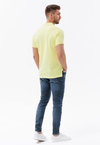 Svetle žluté polo triko s krátkým rukávem a klasickým límečkem, nošené s modrými džíny a bílými teniskami, viděné zezadu.