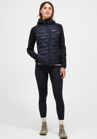 Mörkblå puffväst med quiltad front och enfärgade ärmar, kombinerad med åtsittande svarta leggings och ljusa vandringsskor.