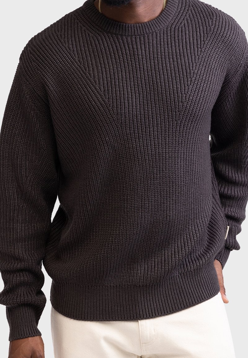 Homme portant un pull en tricot texturé marron foncé avec des poignets et un ourlet côtelés, associé à un pantalon de couleur claire.