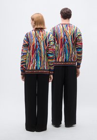 Pull en tricot coloré avec des motifs abstraits en rouge, bleu, vert et jaune, avec un col et des poignets côtelés, associé à un pantalon noir.
