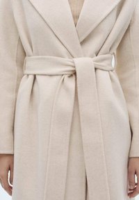 Abrigo envolvente beige hecho de una tela suave con cinturón. Presenta un cuello con muescas y mangas largas, exhibiendo un acabado suave y texturizado.