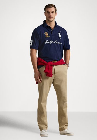 Moški v temno modri polo majici z logotipi, bež hlačah, belih supergah in rdečem puloverju, vezanem okoli pasu, stoji ob enobarvnem ozadju.