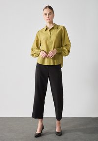 Camicia verde oliva con bottoni, in tessuto testurizzato e maniche voluminose arricciate, abbinata a pantaloni neri a 3/4 e tacchi neri.