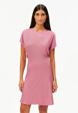 Vrouw met lang donker haar, gekleed in een roze jurk met korte mouwen tot op de knie, staand tegen een effen witte achtergrond.