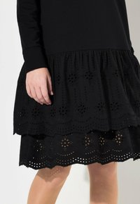 Robe noire avec un corsage texturé et une jupe superposée ornée de broderies et de découpes, accentuée par un ourlet arrondi.