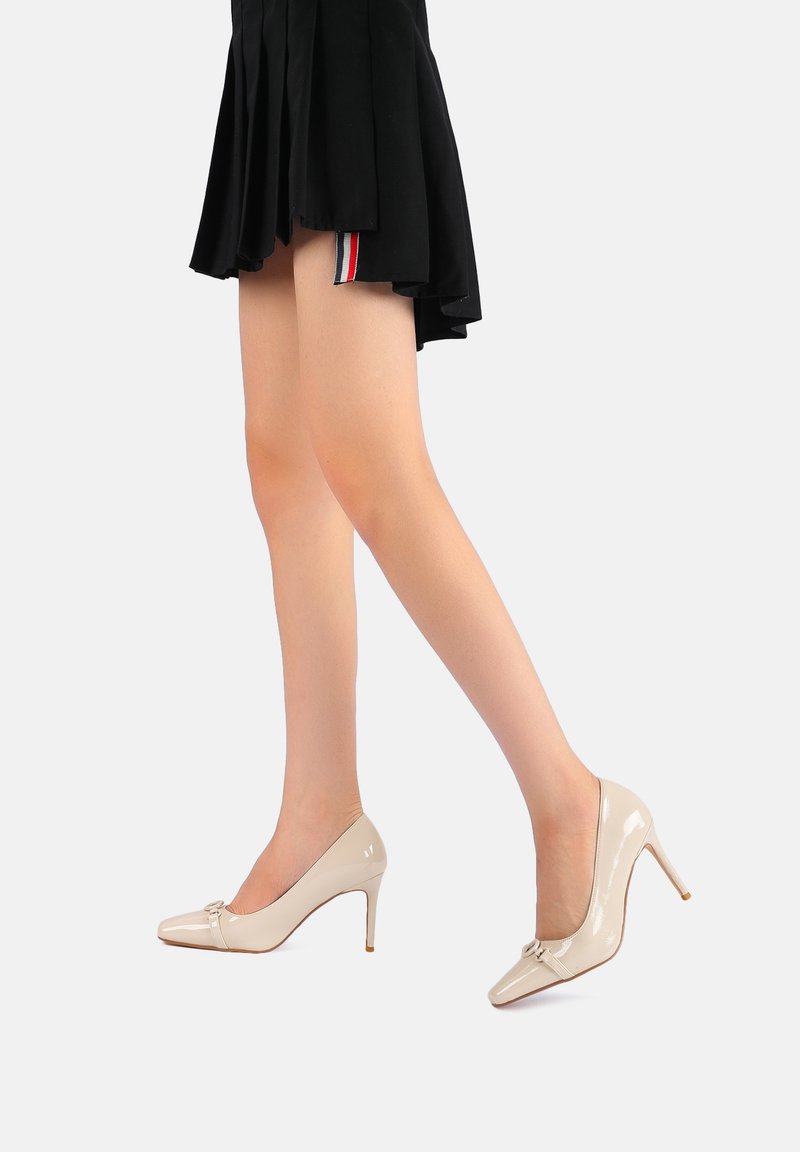 Scarpe con tacco alto in pelle verniciata beige, con punta affusolata e dettagli a fiocco decorativo. Texture liscia, lucentezza sottile e design del tacco slanciato.