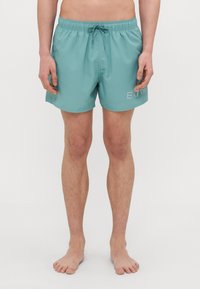 Turkoosi uimapuvut, joissa on naru vyötäröllä, kevyestä materiaalista valmistettu rakenne ja valkoinen logotaittu sivulla. Tyylikäs, polvenpituinen muotoilu.