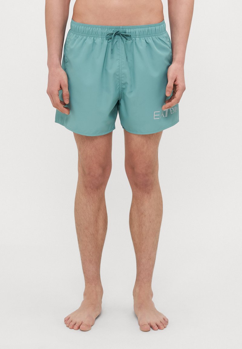 Turkoosi uimapuvut, joissa on naru vyötäröllä, kevyestä materiaalista valmistettu rakenne ja valkoinen logotaittu sivulla. Tyylikäs, polvenpituinen muotoilu.