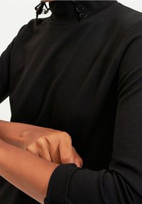 Haut à col roulé noir en tissu doux, avec un détail de bouton sur le col et des manches longues ajustées, au toucher lisse.