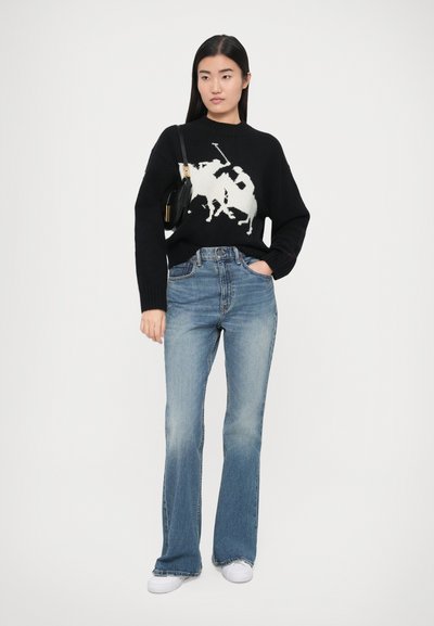 Črn pleten pulover z belo grafiko igralca polota, združen z svetlo modrimi razširjenimi kavbojkami in belimi supergami. Videz je dopolnjen z majhno črno torbico.