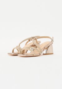 Sandales dorées à talons rétros avec un design à plusieurs brides, accents métalliques et un talon carré. Texture lisse avec une finition brillante.
