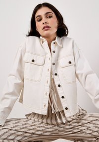 Weiße Jeansjacke mit sichtbaren Nähten, zwei Brusttaschen und Metallknopfverschluss. Über einer gemusterten Bluse mit gebundenem Detail getragen.