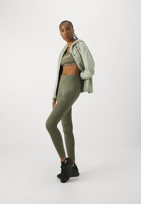 Khakifarbenes Sportoutfit bestehend aus einem gekürzten Oberteil und hochgeschnittenen Leggings. Die enganliegenden Materialien sind glatt und haben dezente Logodetails. Schwarze Sneaker.