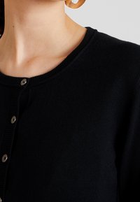 Sort cardigan med ribbet halsudskæring, der har fem sølvfarvede trykknapper og en blød, struktureret strik. Close-up fokuserer på skulderområdet.