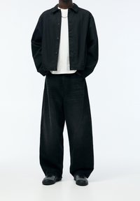 Musta napitettava takki valkoisen t-paidan päällä, yhdistettynä leveälahkeisiin mustiin farkkuihin ja mustiin kenkiin. Yksinkertainen muotoilu, rento istuvuus ja yksiväriset värit.