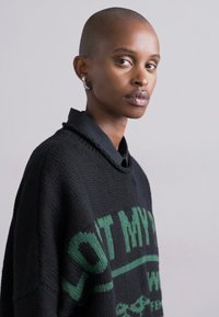 Μαύρος πλεκτός σίντερ με πράσινα γράμματα, με στρογγυλή λαιμόκοψη και χαλαρούς ώμους. Το μοντέλο έχει κοντά μαλλιά και ελάχιστα αξεσουάρ.