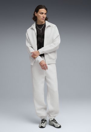 Joven vestido con chándal blanco, camiseta gráfica negra, collar de cadena plateada y zapatillas plateadas, posando frente a un fondo de degradado gris.