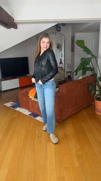 Νεαρή γυναίκα που φορά μαύρο δερμάτινο μπουφάν, μπλε τζιν και μπεζ παπούτσια, στέκεται μέσα σε ένα μοντέρνο σαλόνι με ξύλινο πάτωμα και μεγάλο φυτό.