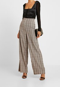 Černý top s dlouhým rukávem a čtvercovým výstřihem, doplněný o kalhoty s vysokým pasem a širokými nohavicemi s károvaným vzorem v béžové a hnědé barvě.