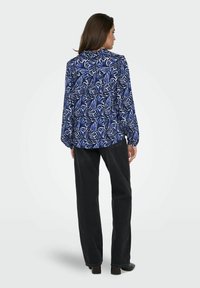 Femme debout, vue de dos, portant un chemisier bleu et blanc à motif de feuilles et un jean noir à jambes larges avec des chaussures noires.