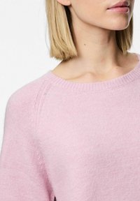 Růžový svetr s měkkou texturou, kulatým výstřihem a raglánovými rukávy. Detaily z blízka ukazují žebrované pletení poblíž výstřihu a švů ramen.