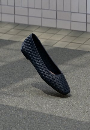 Sort vævet flad sko, der svæver over en asfaltflade med en væg beklædt med fliser i baggrunden.