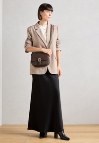 Blazer marron clair, blouse blanche à boutons, jupe maxi noire et bottes à talons noires. Un sac à bandoulière marron et bordeaux avec une boucle en or.