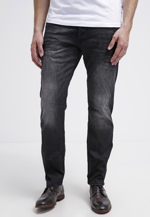 Homme portant un jean slim noir délavé, un t-shirt blanc et des chaussures de ville en cuir marron foncé, debout sur une surface blanche.