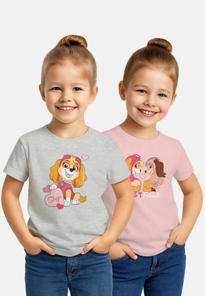 Due ragazze sorridenti con chignon indossano jeans casual e t-shirt con temi di cani animati, in piedi con le mani nelle tasche contro uno sfondo bianco.