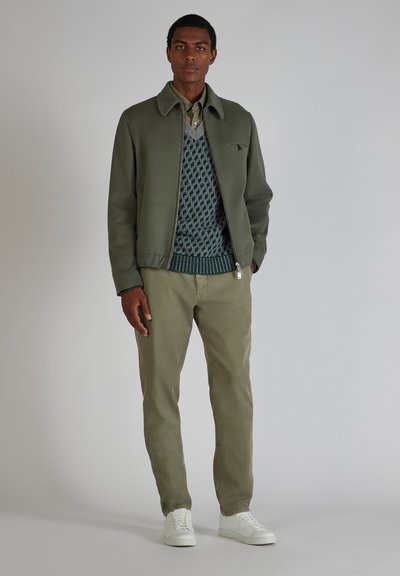 Olivgrüne Bomberjacke über einem blauen strukturierten Pullover mit Rautenmuster, khakifarbene Hose und weiße Sneaker. Schlichtes und lässiges Design.